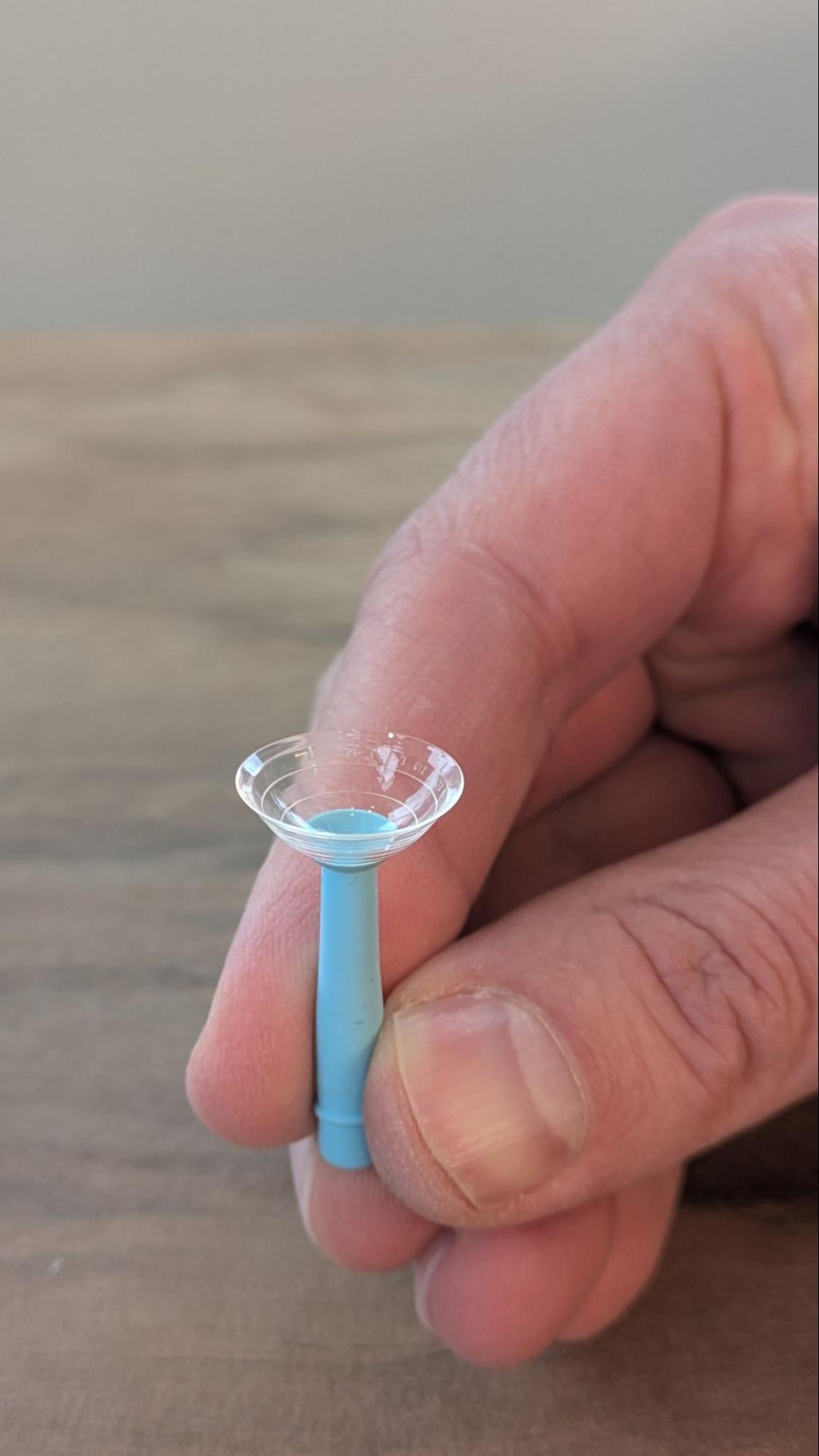 A close-up of a hand holding a filled scleral contact lens on a blue insertion plunger.